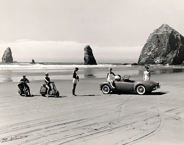 5 young people on the beach at Cannon Beach. 2 sit on scooters. 1 sits on a sports car & holds a beach ball.