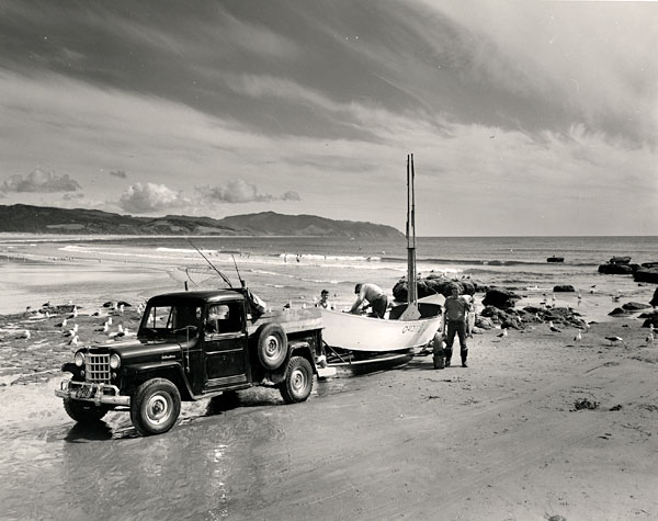 A man stands next to a boat pulled by a Jeep onto the beach. Another man is in the boat bent over and handling gear in the boat.
