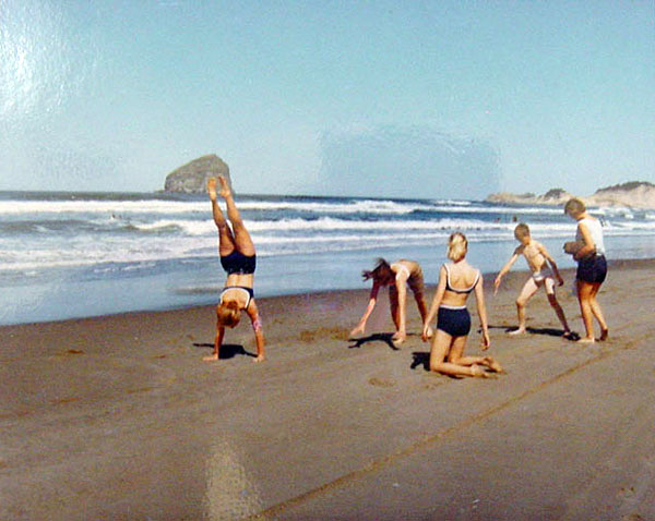 A mother with 3 teen daughters & 1 young boy play on the beach. 1 girl does a handstand at the water's edge.