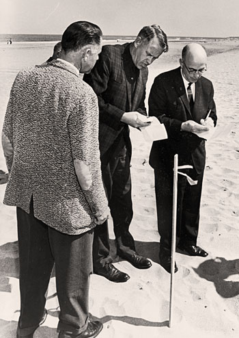 3 men stand next to a stake in the sand. 2 of the men are making notes on paper.