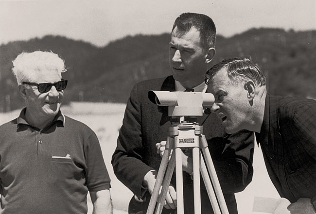 3 men stand on a beach. 1 looks through the view port of a piece of survey equipment.
