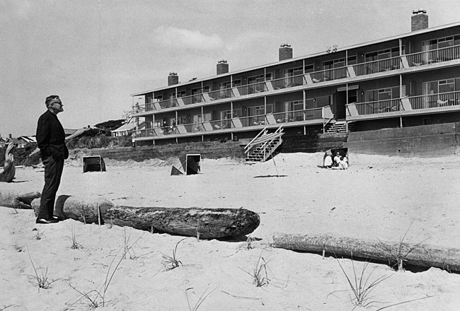 A man stands on the beach by a log looking up the beach toward a motel. Steps lead from the motel porch to the beach.