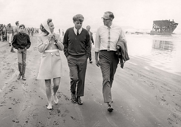 A woman walks with 2 men down the beach. Crowds of people are seen walking behind them. The wreck of a ship is seen in the ocean