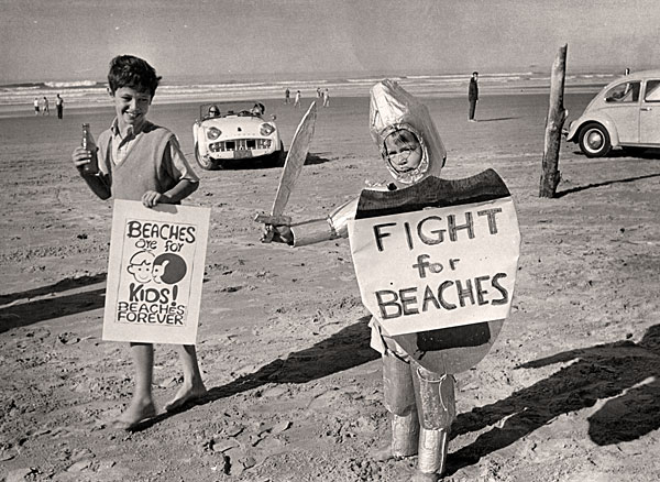 2 boys on the beach with signs. 1 boy dressed as a knight with a cardboard sword. Signs read: "Fight for Beaches" & "Beaches are for kids!"