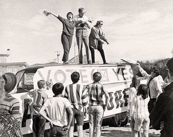 A woman speaks to a crowd with her arms outstretched on top of a van with a sign reading: "Vote 6 yes"