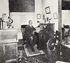 A black man in a suit sits at a desk in the early 1900s