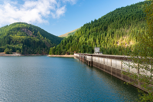 A scenic view of a dam with a large reservoir in the foreground, surrounded by dense forests on rolling hills under a clear blue sky. The dam, made of concrete, spans across the image with water visibly calm and reflecting the surrounding nature.