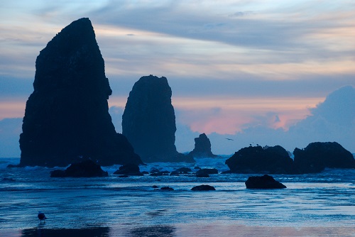 Tall rock formations along the coast line where the water meets sand. The sun sets in the distance.