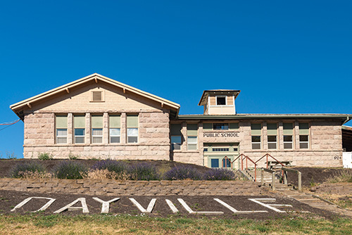 A single-story public school building made of stone blocks with a sign that reads DAYVILLE PUBLIC SCHOOL. In the foreground, the word DAYVILLE is spelled out in large white letters on a grassy slope.