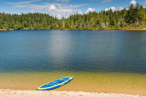 A canoe rests on a sandy beach, part way in the water. The calm lake has fir trees lining the far bank.