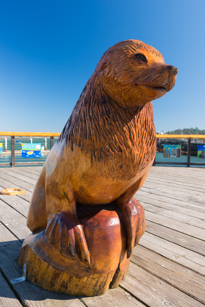 A sea lion carved from a tree sits on a boardwalk near the ocean.