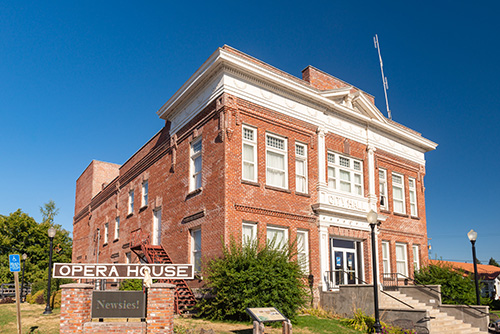 A two-story red brick building with white trim and a sign that reads OPERA HOUSE in capital letters. The building features large windows on the second floor, a decorative cornice, and an American flag on top.