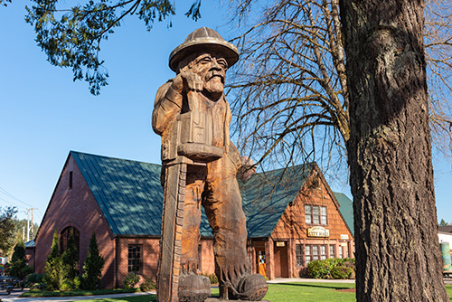 The image depicts a large wooden statue of a person wearing a hat and overalls. The statue stands in the foreground, holding a square sign that covers the face.