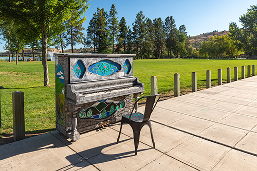An ornately decorated public piano and a single chair placed outdoors on a concrete pavement.