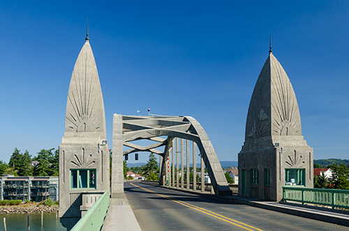 A clear day view of a unique bridge with two large, pointed, art deco-style concrete pylons at the entrance. The bridge features a metal arch supporting the roadway and has green railings on either side. The sky is blue, and there are trees visible in the background.