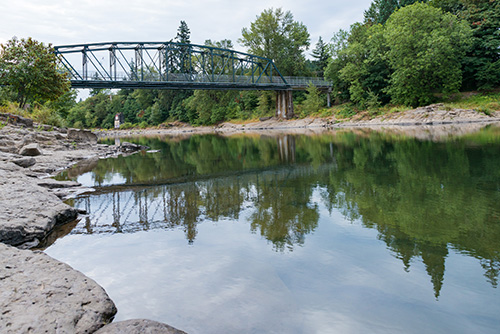 A steel truss bridge spanning over a calm river with reflections on the water surface. The riverbank is lined with rocks in the foreground and trees in the background.