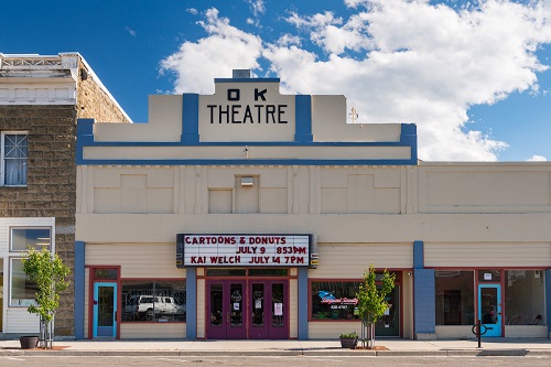 The theater building communicates its function through a prominently display O.K. Theatre name plate in the center of a stepped parapet. The building is constructed of reinforced pour-in-place, board-form concrete.