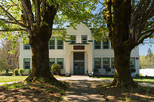 A symmetrical two-story building with a central red door, flanked by two large trees. The building has a white facade with green trim around the windows and roofline. A concrete pathway leads to the entrance, and the grass on either side is well-manicured. 