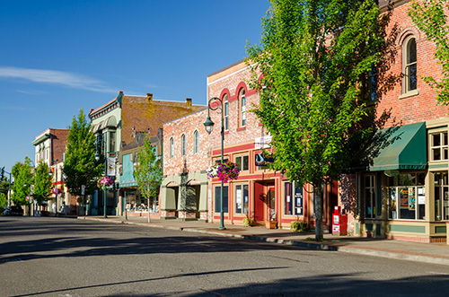 A sunny day on a quaint main street with a clear blue sky. The street is lined with charming, historic two-story buildings featuring various architectural details such as brick facades and decorative trim. The buildings house small businesses including a café with outdoor seating, indicated by the presence of tables and chairs on the sidewalk.