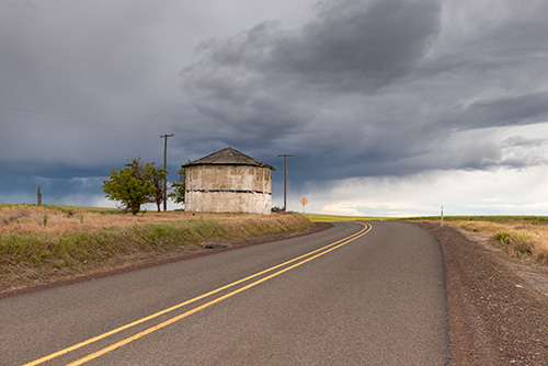 A two-lane road curves to the right leading towards a horizon under a dramatic sky with dark, stormy clouds. On the left side of the road, there is an old, weathered storage building with a peaked roof.