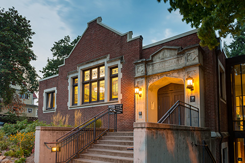 Exterior view of a brick building at dusk with warm lighting emanating from windows and lamps. The building features a classical architectural style with steps leading up to an arched entrance that has Public Library inscribed above the door.