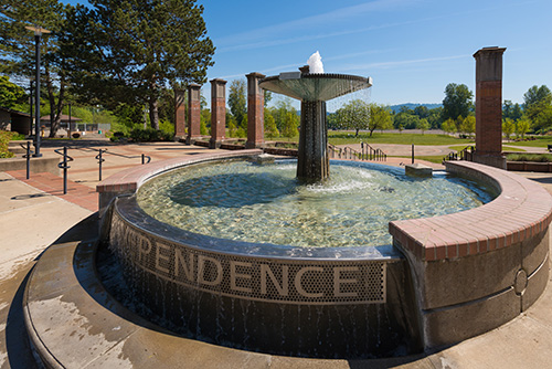 An outdoor circular fountain with water cascading from a tiered central structure. The word INDEPENDENCE is prominently displayed on the side of the fountain in capital letters.