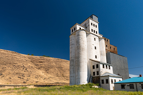 A building constructed of concrete with several small windows scattered across its facade. It is situated in a dry, grassy field with a gentle hill in the background.