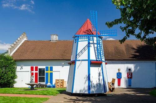 A windmill about the height of a 1-story home stands in front of a building with the Danish & Swedish flags painted on front.
