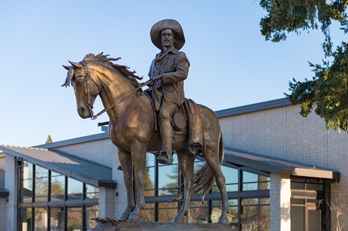 Statue outside on a raised platform of a man in a cowboy hat sitting atop a horse and holding the reins. 