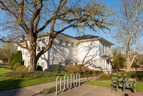 A two-story white building with multiple windows, surrounded by a well-manicured lawn and a large leafless tree.