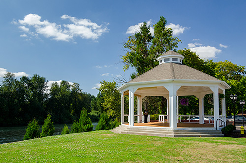 A white gazebo with a hexagonal structure and a cupola on top is situated on a well-manicured lawn beside a body of water.