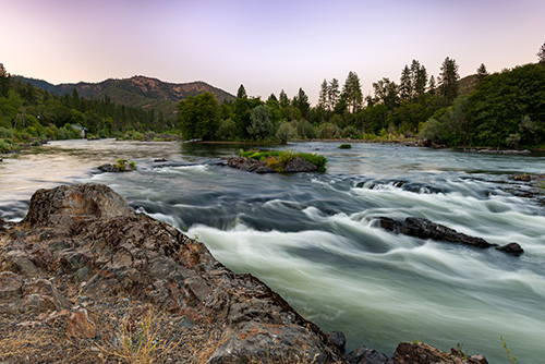 Landscape featuring a river with rapid flowing water, surrounded by lush greenery and trees. In the foreground, there’s a rocky outcrop overlooking the river.