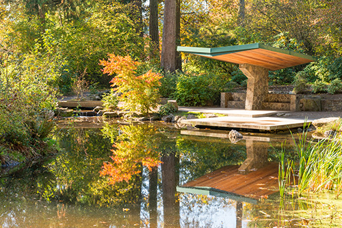 Outdoor scene with a calm pond reflecting the surrounding nature. A wooden dock extends over the water, topped with a green-roofed shelter.