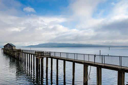 A long wooden pier extending into a calm body of water with overcast skies. The horizon is obscured by low-lying clouds, and distant landforms are faintly visible through the mist.