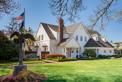 A two-story building with white siding and a dark shingle roof. The building has multiple windows, a chimney, and an American flag on a pole to the left.