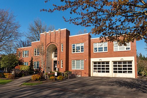 A two-story brick building with a prominent arched entrance and multiple white-framed windows. There are two garage doors on the ground level, suggesting it might be a fire station or municipal facility.