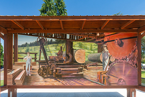 An outdoor depicts a historical logging scene with green hills in the background. On the left, an individual wearing a hat and long-sleeve shirt stands beside a large steam donkey engine used for logging.