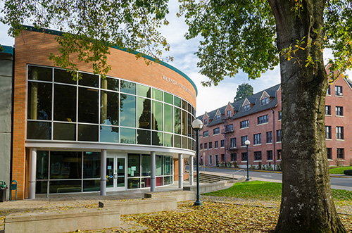 Exterior view of a modern building with a curved facade featuring large glass windows. The building is adjacent to a traditional brick building with dormer windows. A tree with green leaves is visible on the right, and the scene is set against a backdrop of a clear sky with few clouds.