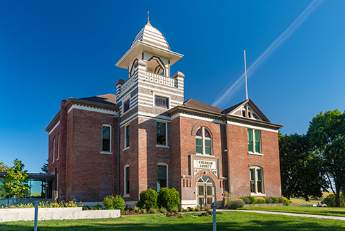 A historic red brick building with white trim and a distinctive tower under a clear blue sky, labeled Buchanan County Courthouse. The building is surrounded by well-manicured green lawns.