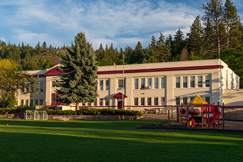 A two-story school building with a red roof and trim, surrounded by lush greenery under a clear blue sky. The foreground shows a well-maintained grassy field with playground equipment and soccer goals.