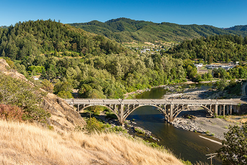 A scenic view of a concrete arch bridge spanning across a river, surrounded by lush greenery and hills under a clear blue sky. Dry grass in the foreground contrasts with the verdant landscape, highlighting the region’s diverse vegetation.