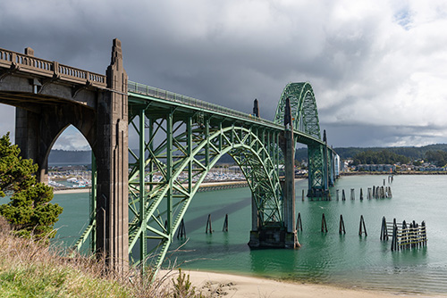 A large green steel arch bridge spans across a body of water with a cloudy sky overhead. The bridge features intricate trusses and is flanked by concrete towers at its entrance, with remnants of old piers in the water.