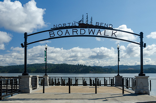 An ornate metal archway reading North Bend BOARDWALK stands at the entrance to a wooden boardwalk, with two lamp posts on either side. In the background, a serene lake and mountain range under a partly cloudy sky.