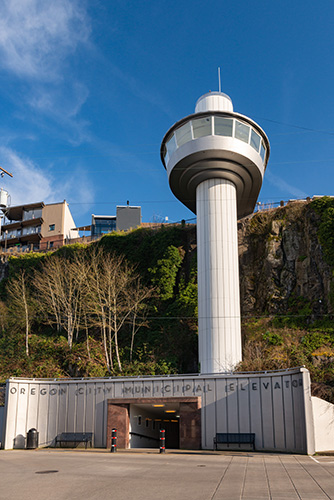 A tall, cylindrical observation tower with a circular viewing platform and a smaller upper section stands against a clear blue sky. Below the tower is the entrance to the Oregon City Municipal Elevator, built into a natural cliff face with large letters spelling out its name.