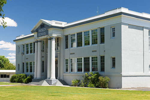 A classic white school building with a sign reading PAISLEY SCHOOL above the entrance, which is flanked by four large columns. The school is set against a clear blue sky and surrounded by green lawns.