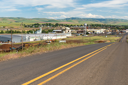 A paved road leads towards a small industrial town with several large white storage tanks and buildings. In the background, rolling green hills under a blue sky with scattered clouds.