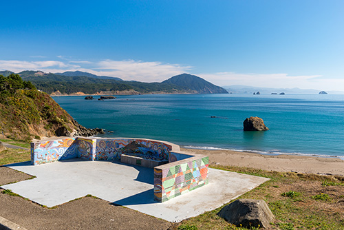 A scenic coastal landscape with a clear blue sky. In the foreground, a concrete platform with colorful graffiti overlooks a calm sea, with hills, mountains, and small islands visible in the distance.