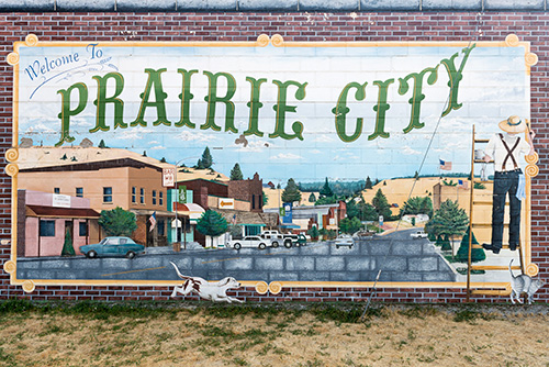 A mural on a brick wall depicting the words Welcome to PRAIRIE CITY in large, stylized green letters. Below the text is a painted scene of a small town with buildings, cars, and distant mountains under a blue sky.