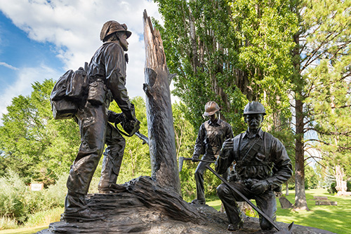 A bronze statue of two firefighters stands in a park, one holding a hose and the other kneeling, with trees and a blue sky in the background.