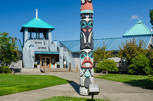 A vibrant totem pole stands in the foreground with intricate carvings and colorful patterns, featuring faces and symbols. In the background, a building with a blue roof and a gazebo-like structure is visible under a clear blue sky.